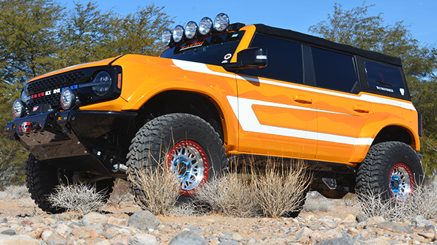 orange Ford Bronco with white stripes and light bra and red white and blue logo and MetalCloak front skidplate on dirt gravel path near trees